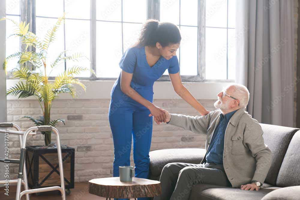 Smiling caring woman doctor talk consult satisfied elderly male patient, give visit to retired grandfather at home, happy attentive female nurse do regular checkup consultation to mature man client